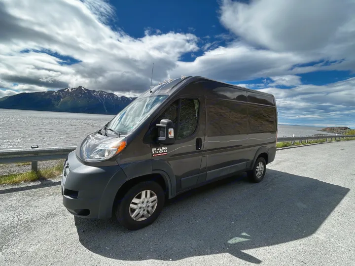 Dark grey Ram ProMaster RV rental parked on a scenic road by mountains and water under a cloudy sky, ideal for adventure.. Dodge Ram 1500 2017