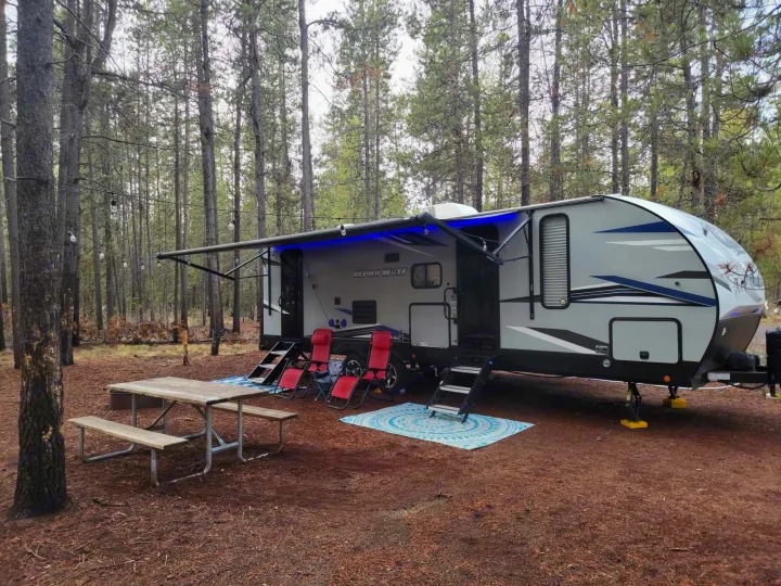 This inviting RV rental campsite features a travel trailer with an extended awning, blue lights, and a picnic area in a pine forest.. Forest River Cherokee Alpha Wolf 2021