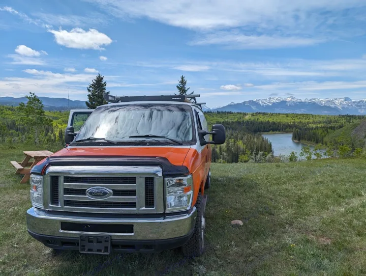 Orange RV rental van offers stunning views of a serene lake, lush forests, and majestic snow-capped mountains from a grassy hill.. Ford Econoline E250supercab 2012