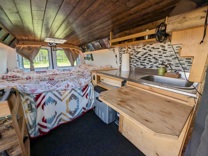 Cozy RV rental camper van interior featuring a comfy bed with colorful blankets, a wood ceiling, and a compact kitchen area with a sink.. Ford Econoline E250supercab 2012