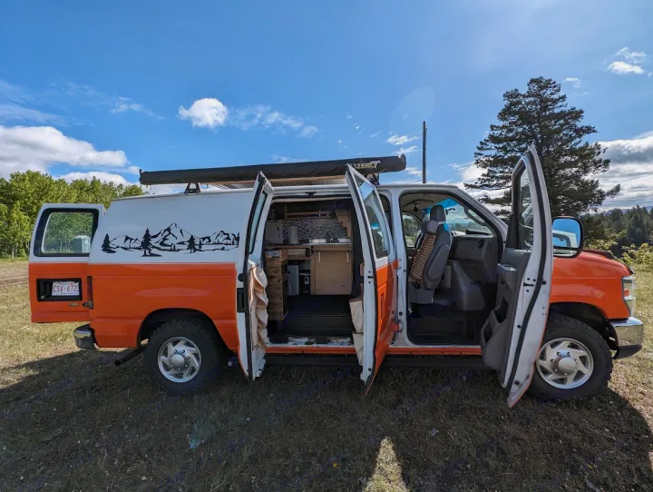 This customized RV rental van, with open doors revealing its interior and mountain decal, is ready for adventure in the outdoors.. Ford Econoline E250supercab 2012