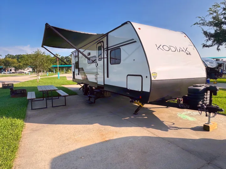 White KODIAK SE travel trailer RV rental at a sunny campsite, awning extended, with a picnic table & fire pit.. Dutchmen Kodiak SE 2024