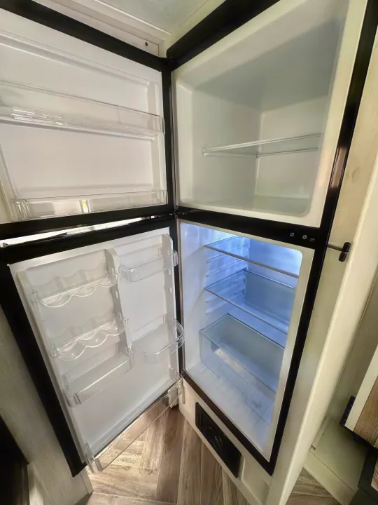 Empty, sparkling clean refrigerator in an RV rental, showing clear shelving and door bins, ready for use.. Dutchmen Coleman Rubicon 2023