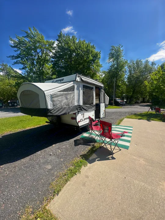 Relaxing at a campsite with a white pop-up RV rental, red chairs on a striped mat, and green trees under a blue sky.. coachmen clipper 2018