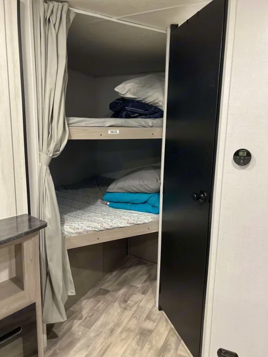 An RV rental's sleeping area with two neatly made bunk beds, a privacy curtain, and a digital thermostat visible.. Dutchmen Kodiak SE 2024