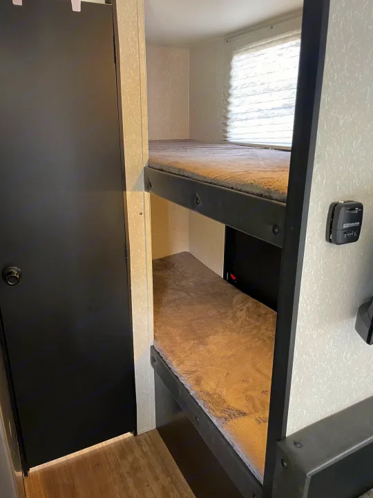 Interior of an RV rental showing cozy bunk beds with fuzzy blankets, a bright window, and a dark door on the left.. Forest River Cherokee Wolf Pup 2022