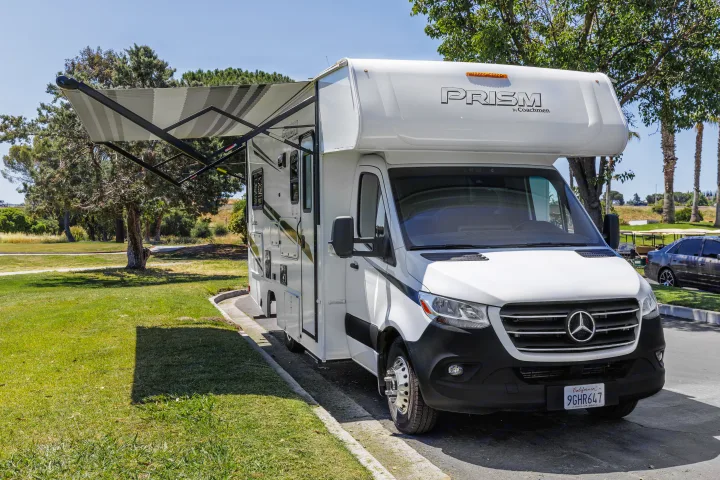 Pristine white RV rental (Coachmen Prism) with extended awning, parked on a sunny day next to a lush green park.. Coachmen Prism 2023