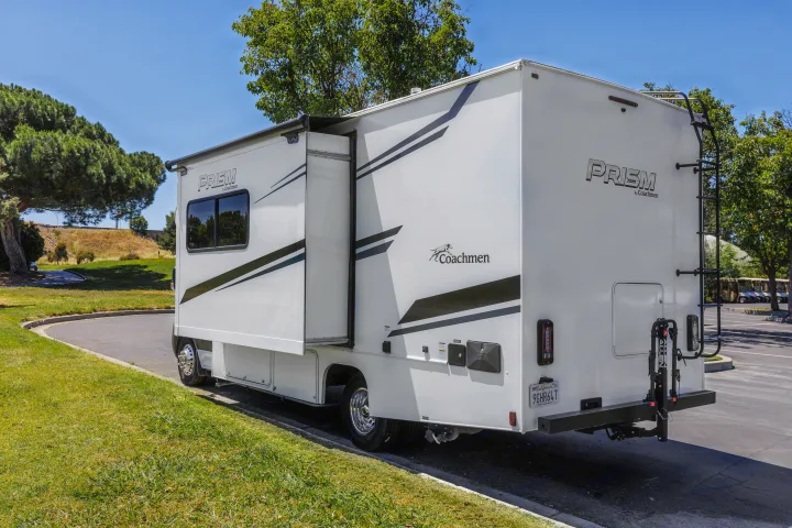 A white Coachmen Prism RV rental with extended slide-out, ladder, and bike rack. Parked for camping.. Coachmen Prism 2023