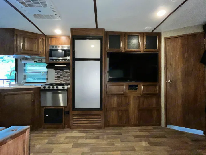 Inside an RV rental: a modern kitchen with stove, microwave & fridge, plus living area with TV, all in warm wood tones.. Keystone RV Cougar X-Lite 2018