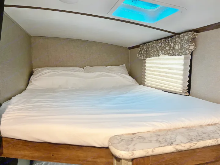 Step into comfort in this RV rental's bedroom. Features a large bed with white linens, window, and bright overhead skylight.. Keystone RV Cougar X-Lite 2018