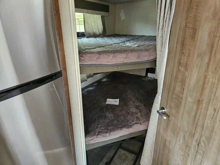 Inside an RV rental: two cozy bunk beds with quilted mattresses, curtains, and a fridge on the left.. Forest River Flagstaff Micro Lite 2023