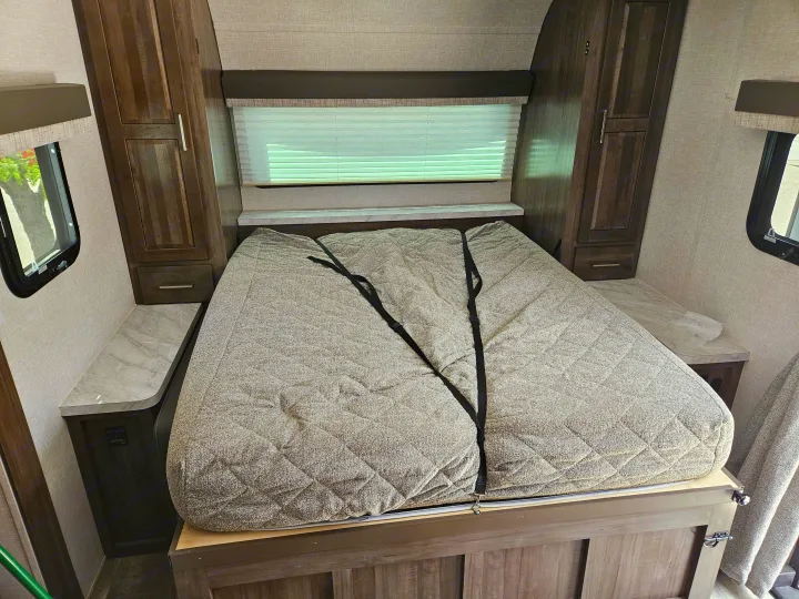 Clean RV rental bedroom showing a gray quilted mattress secured by straps, dark wood cabinets, and windows with blinds.. Forest River Flagstaff Micro Lite 2023
