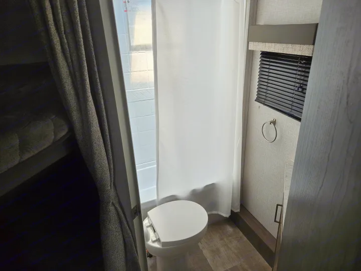 Compact RV rental bathroom with toilet, white shower curtain, and bunk bed visible on the left. Ideal for small spaces.. Forest River Flagstaff Micro Lite 2023
