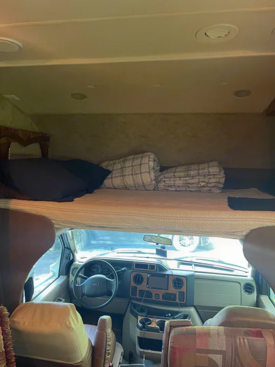 Inside an RV rental, showing the front cockpit with steering wheel and dash, and an overhead bunk bed with bedding.. Jayco Greyhawk 2010