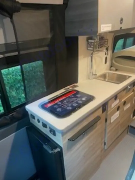 Inside an RV rental, a compact kitchenette features a sink, faucet, fridge below, and wooden storage drawers, ideal for travel.. Winnebago Solis 2023