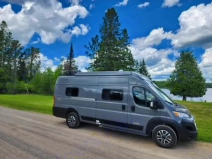 Explore scenic routes! This grey camper van RV rental is parked by a lake under a cloudy sky, ready for your next road trip.. Winnebago Solis 2023