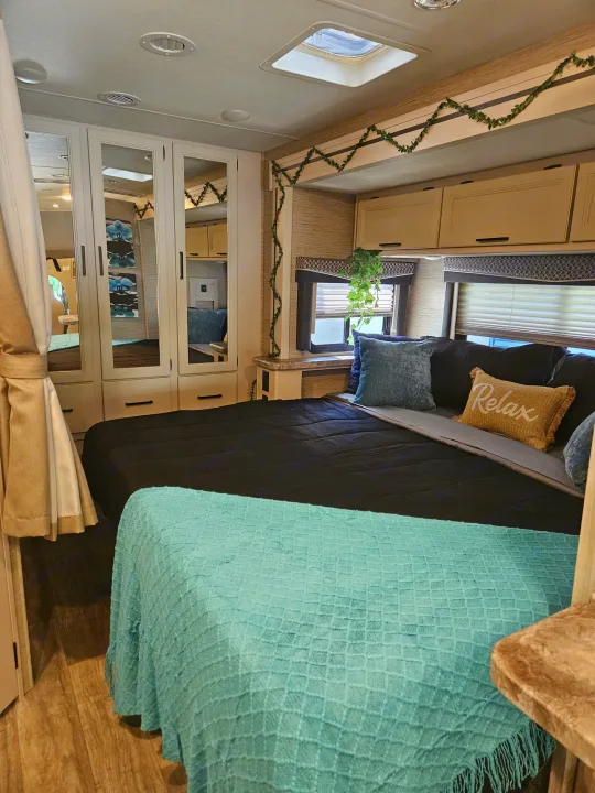 Cozy RV rental bedroom with black bed, blue blanket, mirrored wardrobe, overhead cabinets, and windows, ideal for travel.. Thor Chateau 2023