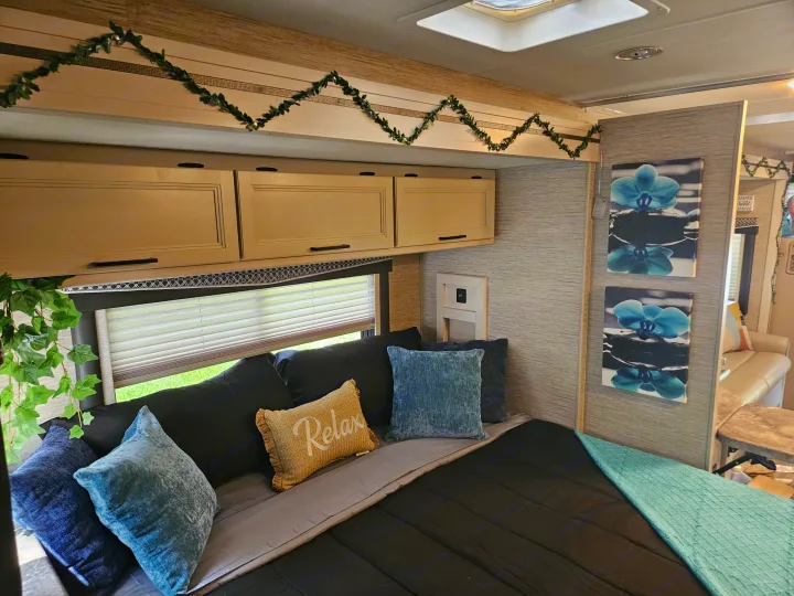 Inviting RV rental bedroom: black bedding, blue pillows (one says "Relax"), overhead cabinets, and orchid art.. Thor Chateau 2023