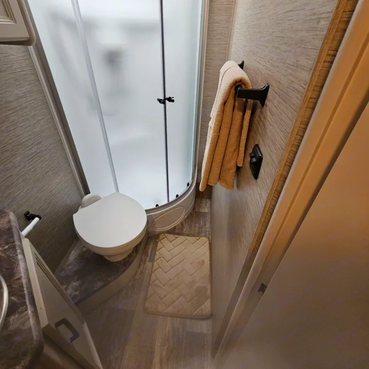 This compact RV rental bathroom features a frosted corner shower, toilet, and convenient towel rack.. Thor Chateau 2023