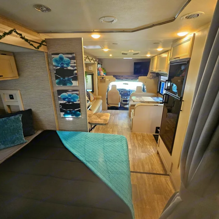 View inside an RV rental: from a bed with teal bedding, looking forward through the living space, kitchen, and driver's area.. Thor Chateau 2023