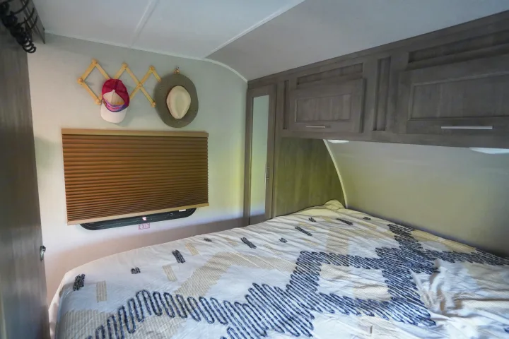 An RV rental bedroom featuring a bed, overhead cabinets, window with blind & exit sign, plus a hat rack.. Forest River Vibe 2020