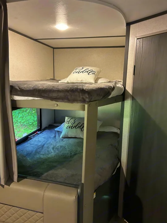 This RV rental features cozy bunk beds, with soft grey blankets, white pillows, and a window offering a view of green trees.. Dutchmen Aspen Trail LE 2022