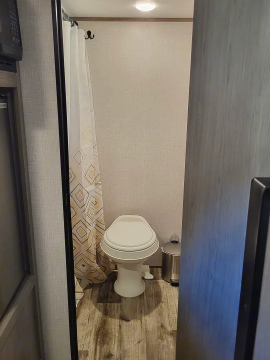 View into a small RV rental bathroom with a white toilet, patterned shower curtain, and light wood-look floor.. Dutchmen Coleman Lantern LT 2023
