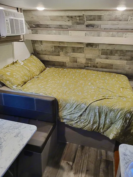 RV rental interior featuring a cozy bedroom area with green floral bedding, mounted AC unit, wood paneling, and dinette.. Dutchmen Coleman Lantern LT 2023