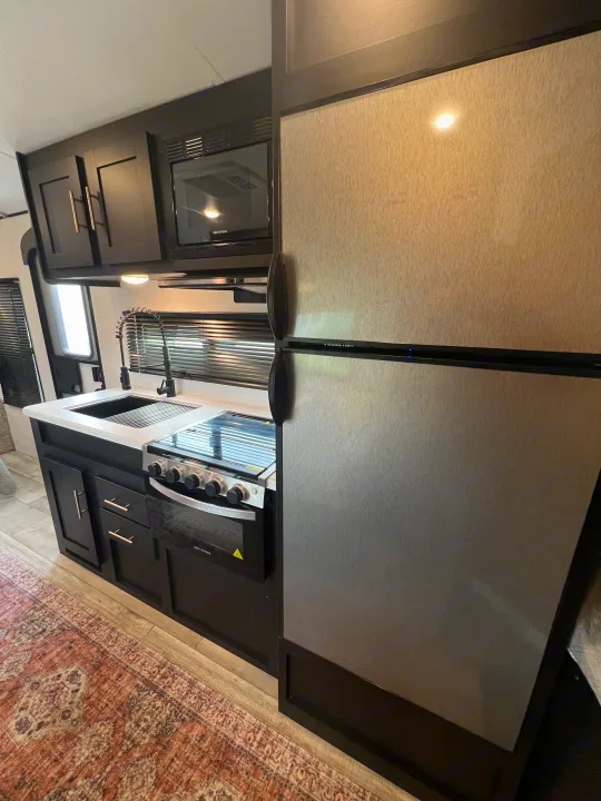 This modern RV rental kitchen showcases dark cabinetry, a brushed metal refrigerator, microwave, stove, and a sleek sink.. Keystone RV Springdale 2022