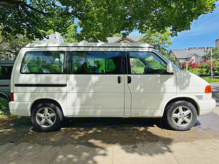 White VW T4 camper van, a perfect RV rental option. Parked on a sunny street under large green trees, residential area visible.. Volkswagen Westfalia 2003