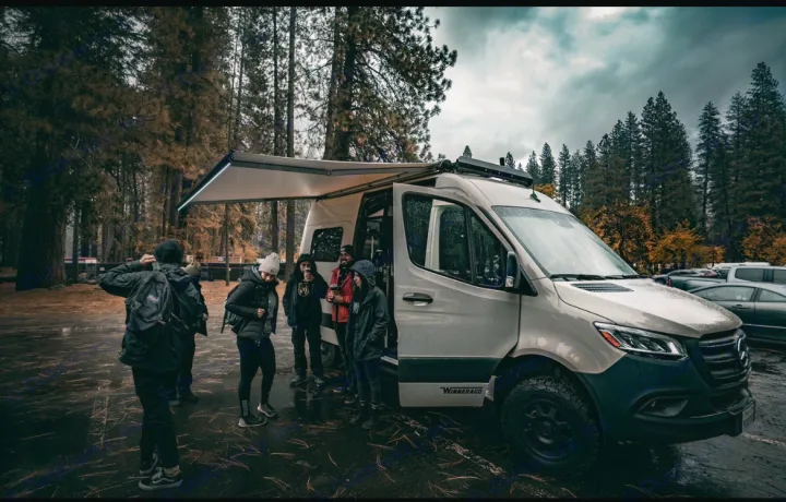 Group enjoys an RV rental van adventure in a damp forest. A light-colored Winnebago with an awning out, surrounded by tall trees.. Winnebago Revel 2022