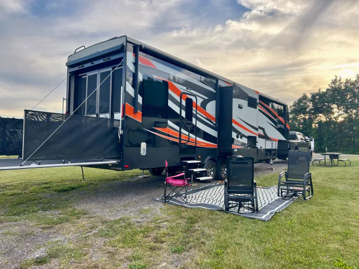 A stylish black, orange, and white toy hauler RV rental is set up at a grassy campsite with an open rear deck and outdoor chairs.. Raptor Raptor 2017