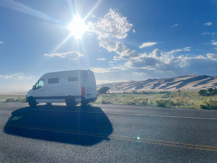 Experience an rv rental road trip! White camper van by majestic sand dunes & road, illuminated by a brilliant sun flare.. Mercedes-Benz Sprinter-Loaded 2022