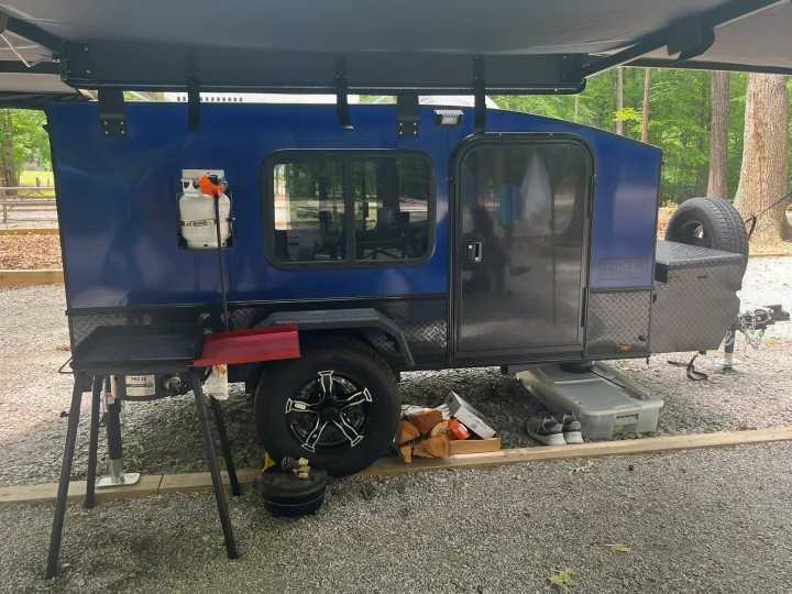 Blue camper trailer with an extended awning, propane tank, and outdoor cooking setup at a gravel campsite. Great for rv rental adventures.. Hiker Trailer highway Deluxe 2023