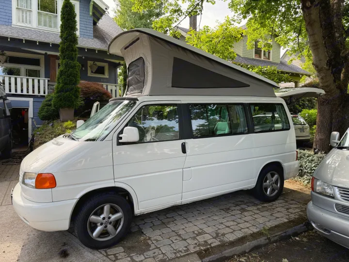 White VW Eurovan pop-top camper, an RV rental parked on a charming cobblestone drive beside a blue house.. Volkswagen Westfalia 2003