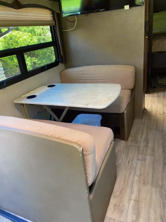Inside an RV rental: a compact dinette with table, upholstered benches, and window overlooking trees, plus TV.. Thor Hurricane 2018
