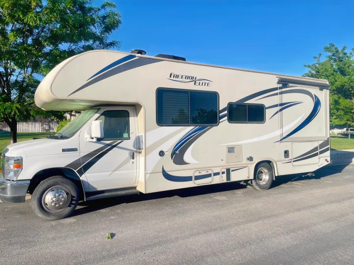 A cream-colored Freedom Elite Class C rv rental, detailed with blue and black graphics, parked outdoors on a sunny day.. Thor Freedom Elite 2019