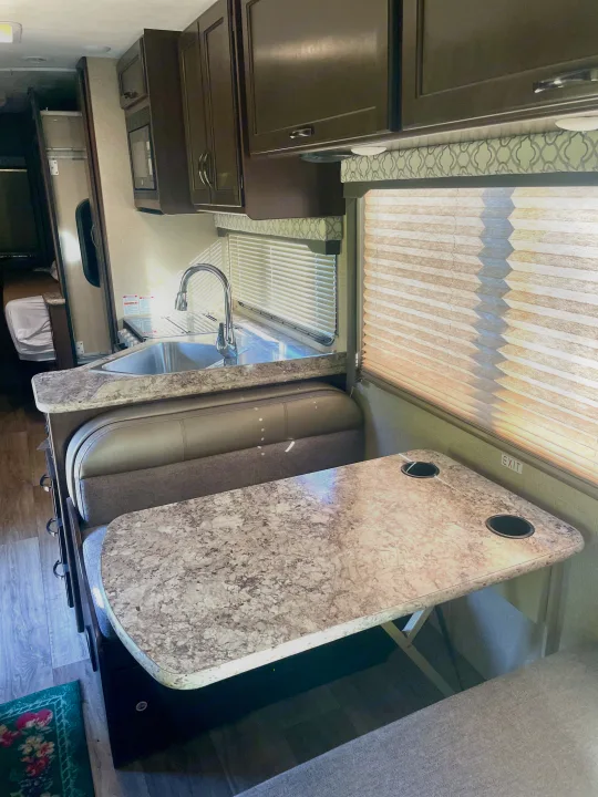 Inside a bright rv rental: functional kitchenette with sink, dark cabinets, and a dinette area with table, seating, & window blinds.. Thor Freedom Elite 2019