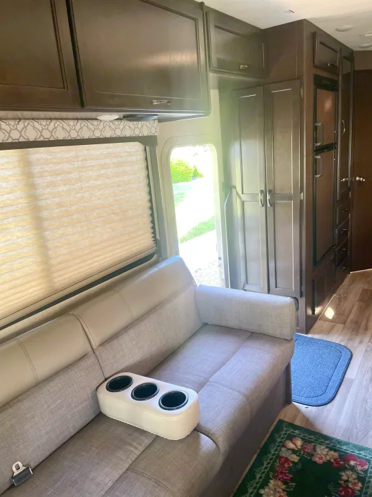 Cozy rv rental interior with a light sofa, integrated cup holders, dark wood cabinets, and an open door to nature.. Thor Freedom Elite 2019
