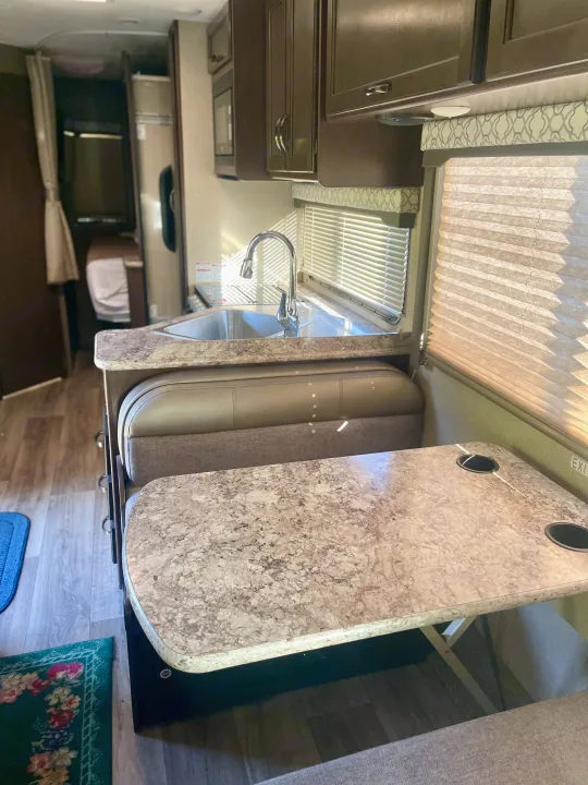 Inside this comfy RV rental: a kitchen with sink, marble-look counter, and dinette with table (cupholders) and window blinds.. Thor Freedom Elite 2019
