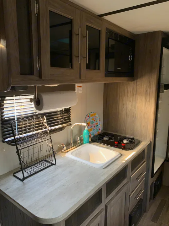 Compact RV rental kitchen with sink, stove, microwave, and wood-grain cabinets, ideal for travel adventures.. Zinger BH 18 2021