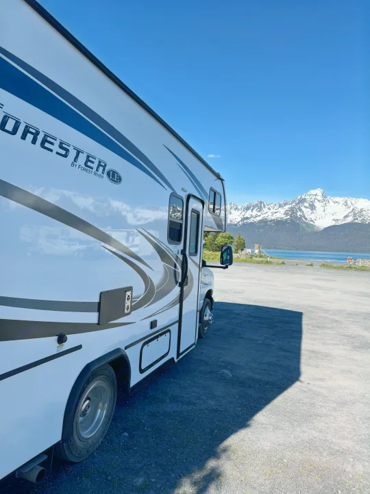 An RV rental sits on a gravel lot, overlooking a serene lake and snow-capped mountains on a clear day.. Forester LE 2022