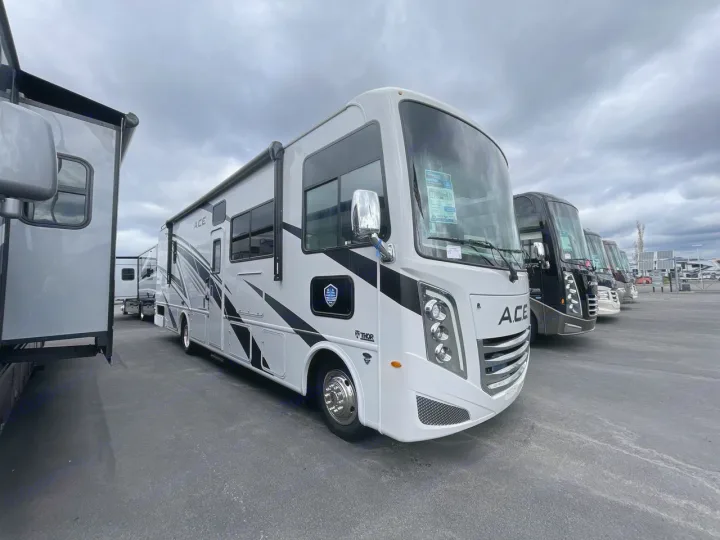 Bright white Thor ACE motorhome at an RV rental and sales lot. Several large RVs are lined up on asphalt under a cloudy sky.. Thor ACE 2024