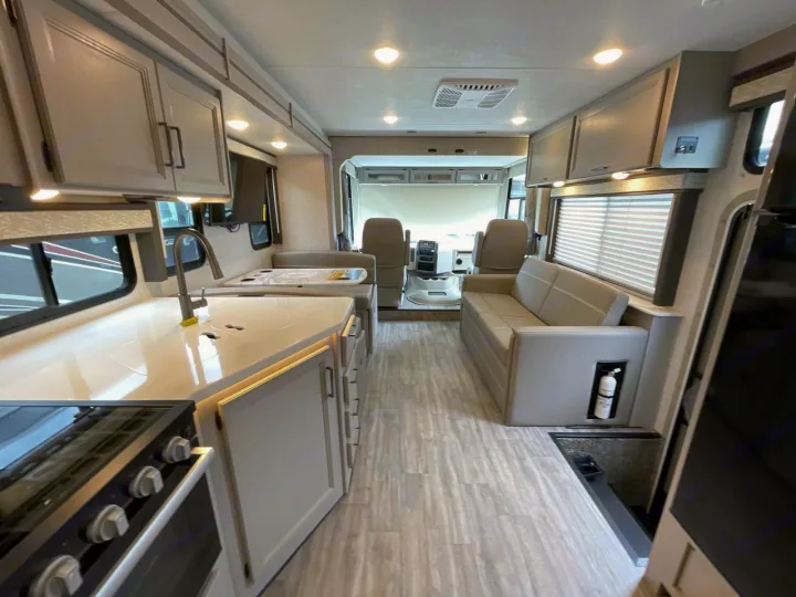 Bright RV rental interior: kitchen with stove/sink, dinette, sofa, and cockpit view. Modern grey cabinets & light wood floor.. Thor ACE 2024
