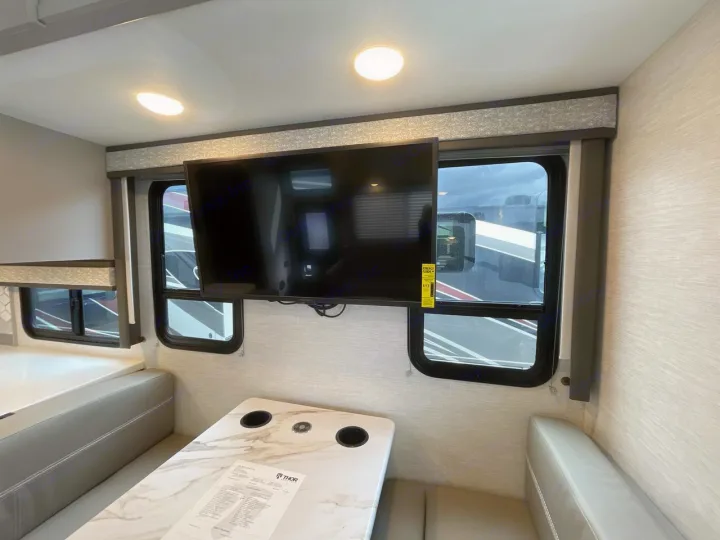 Inside an RV rental: a bright, modern dinette with light benches, a table with cup holders, and a mounted TV, ready for travel.. Thor ACE 2024