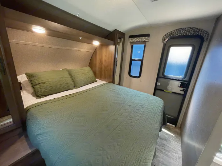 Cozy RV rental bedroom with a queen bed, green quilt, overhead lights, a window, and an entrance door.. Connect 261RL 2018