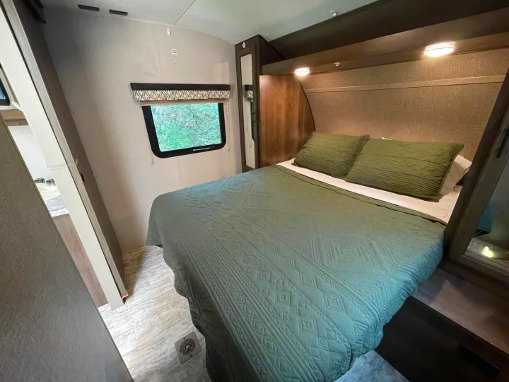 Inside an RV rental, a cozy bedroom featuring a bed with a green quilted comforter, a window to foliage, and a mirror.. Connect 261RL 2018