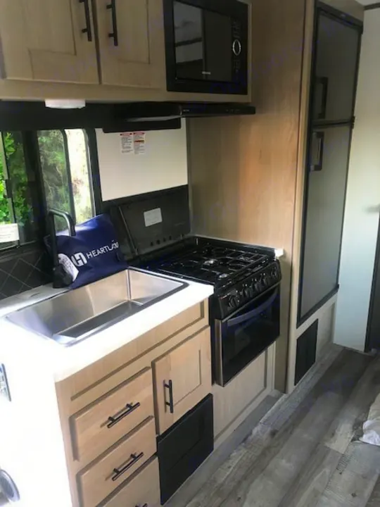 Modern RV rental kitchen with light wood cabinets, stainless steel sink, gas stove, microwave, and refrigerator.. Heartland RVs Other 2022