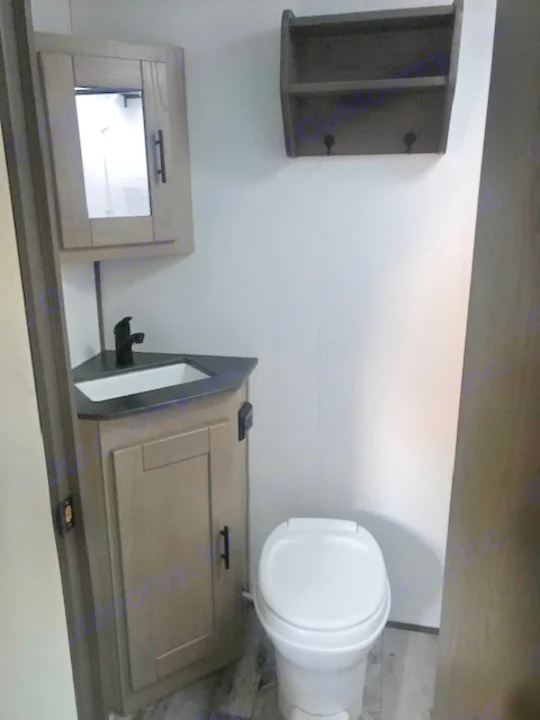 Compact RV rental bathroom with a toilet, corner sink vanity, mirror, and wall shelf for storage.. Heartland RVs Other 2022