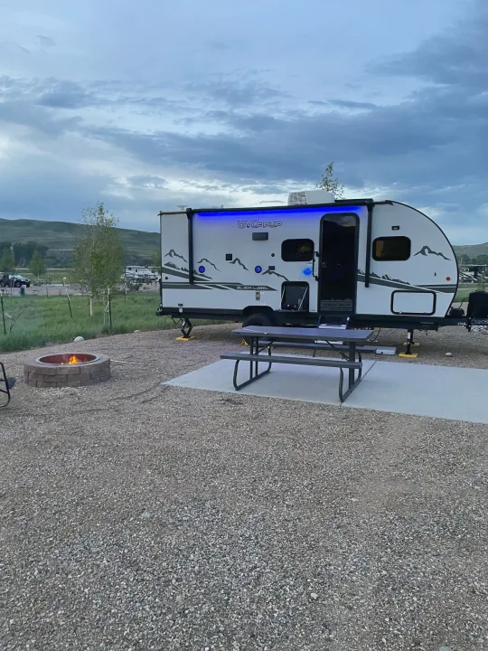 An RV rental, a travel trailer with blue lights, parked at a gravel campsite with a fire pit and picnic table in the evening.. Forest River Cherokee Wolf Pup Black Label 2022